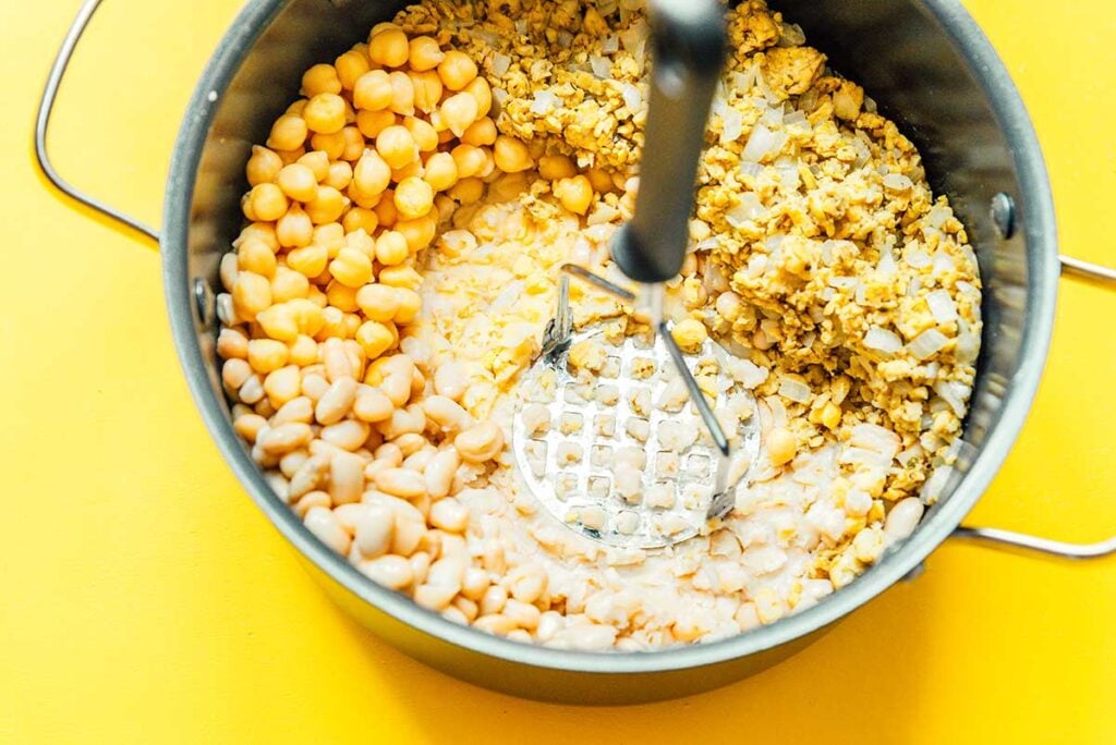 A potato masher mashing the beans in a pot filled with white bean chili ingredients