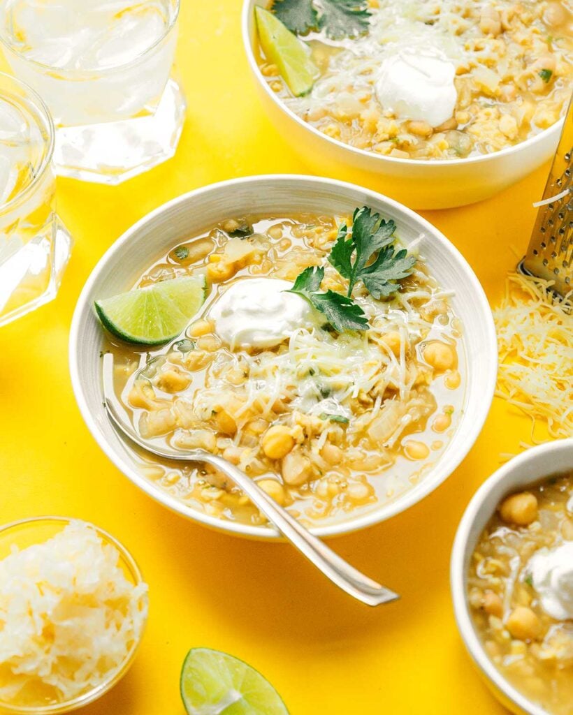 A white bowl filled with tempeh white bean chili and garnished with cilantro, Greek yogurt, shredded cheese, and a slice of lime