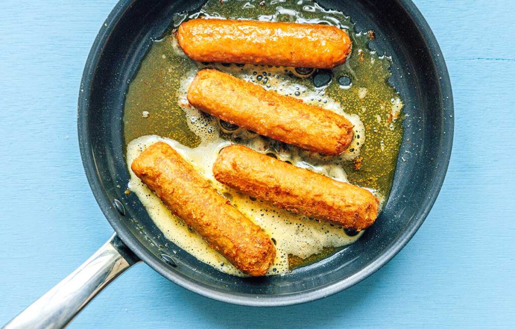 Four plant-based bratwursts cooking in a skillet