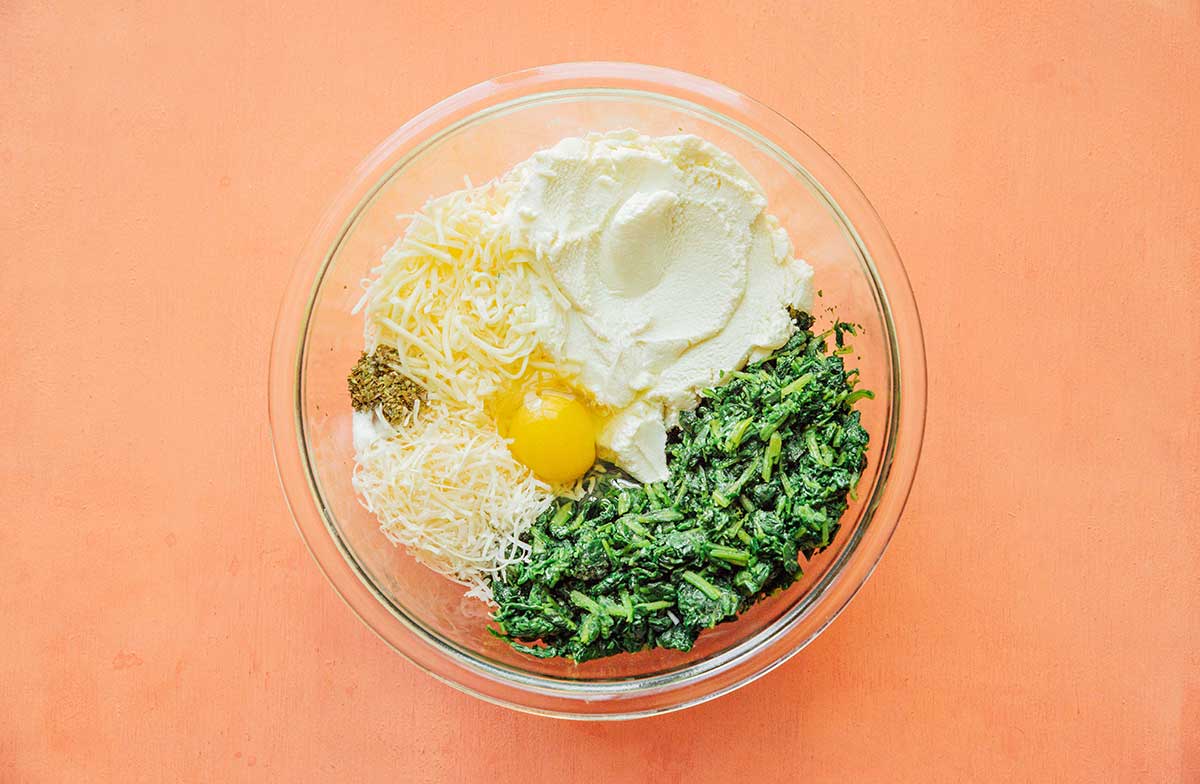 A clear glass bowl filled with the ricotta mixture ingredients including mozzarella cheese, an egg, salt, pepper, oregano, spinach, and parmesan