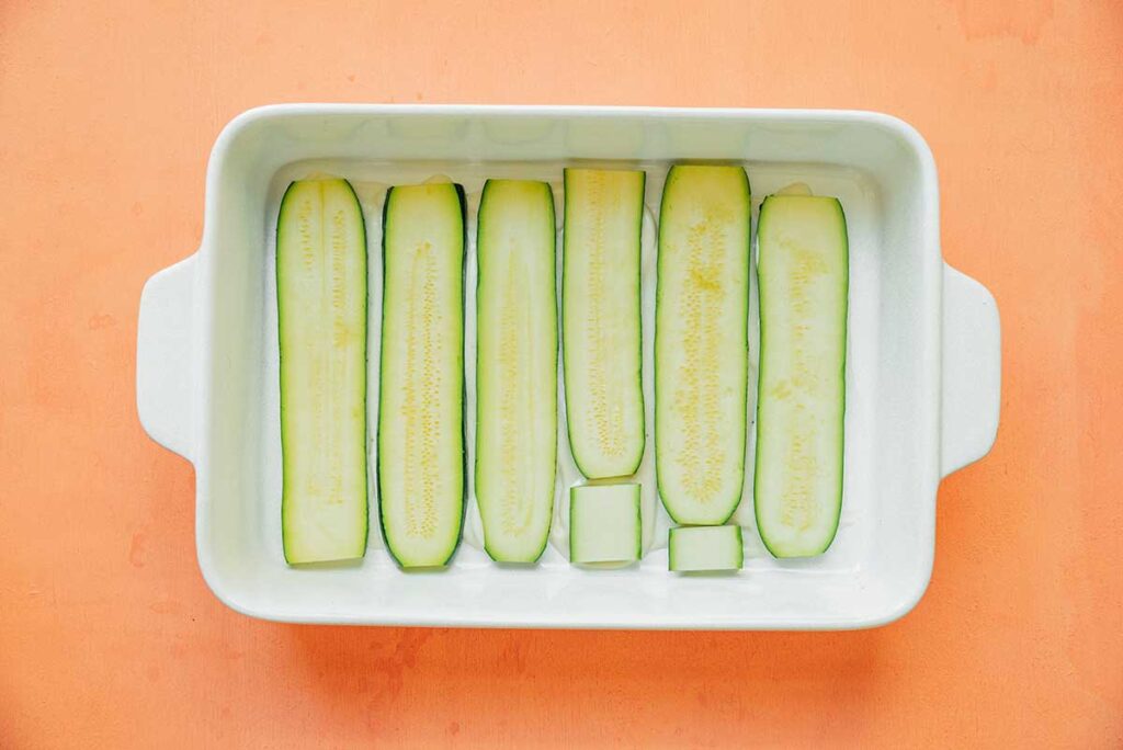 A casserole dish filled with a layer of alfredo sauce and sliced zucchini