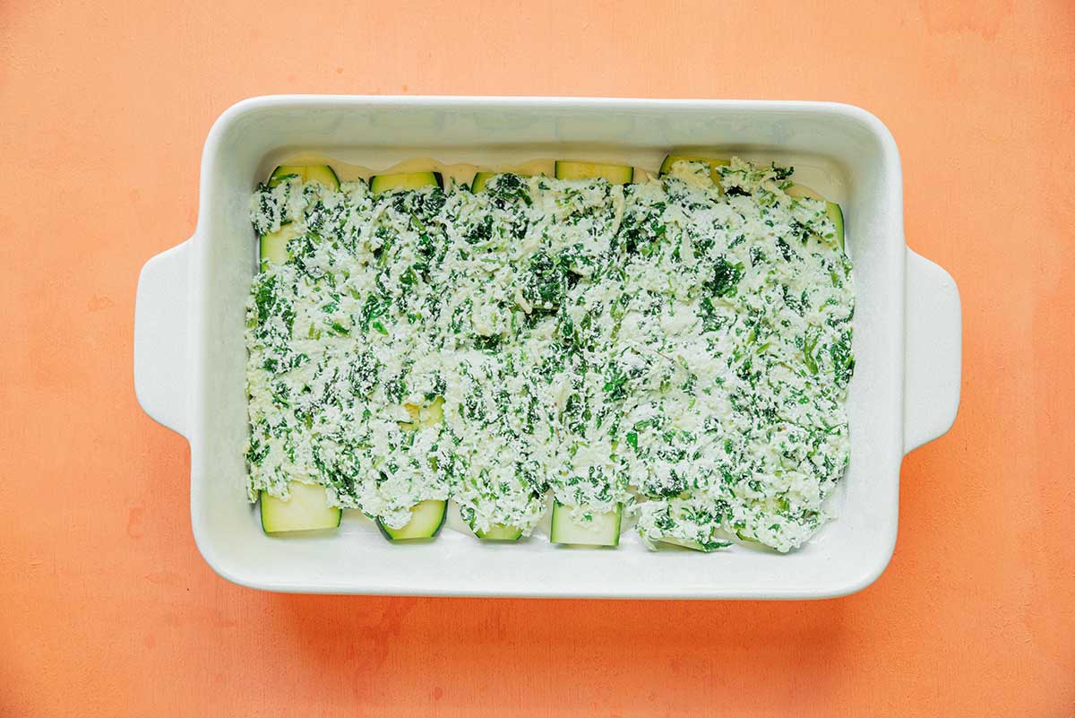 A casserole dish filled with a layer of alfredo sauce, zucchini and ricotta mixture