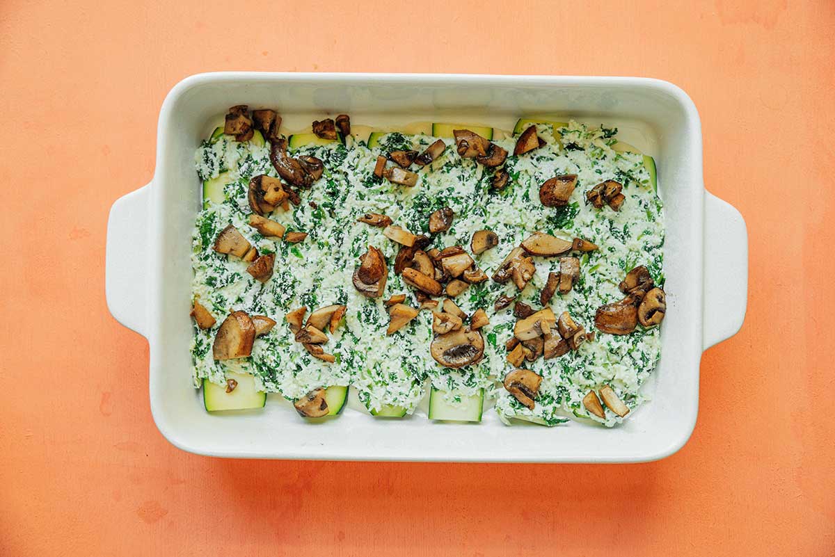 A casserole dish filled with a layer of alfredo sauce, zucchini, ricotta mixture, and sautéed mushrooms