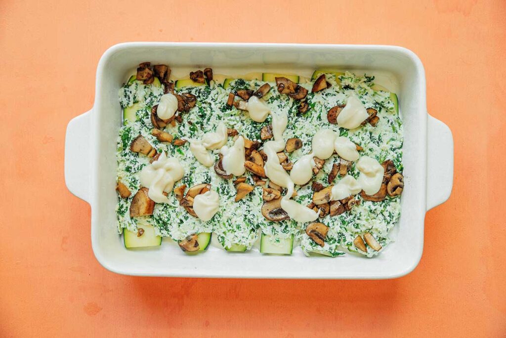 A casserole dish filled with a layer of zucchini, ricotta mixture, sautéed mushrooms, and alfredo sauce