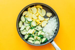 A pan filled with chopped zucchini, yellow squash, onions, and minced garlic sits on a yellow background, reminiscent of the beginnings of a delightful zucchini stew.