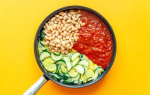 A frying pan filled with a delightful zucchini stew featuring sautéed zucchini, white beans, and rich tomato sauce set against a vibrant yellow background.