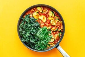 A pan with a delightful zucchini stew brimming with mixed vegetables like beans, tomatoes, and spinach sits invitingly on a yellow background.