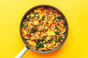A black pan filled with a vibrant zucchini stew, brimming with a colorful mix of chopped vegetables, beans, and greens, set against a bright yellow background.