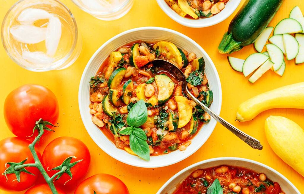 A while bowl filled with zucchini stew and surrounded by ingredients like tomatoes, zucchini, and squash