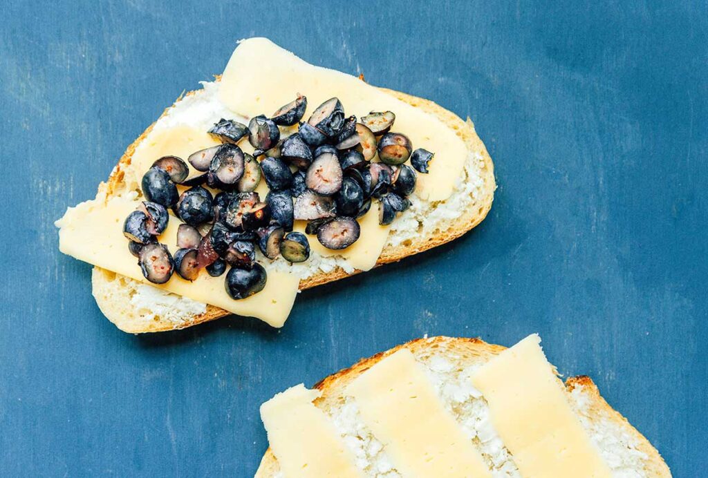 One piece of sourdough bread topped with goat cheese, a slice of mozzarella, and halved blueberries beside a second piece of bread topped with goat cheese and mozzarella