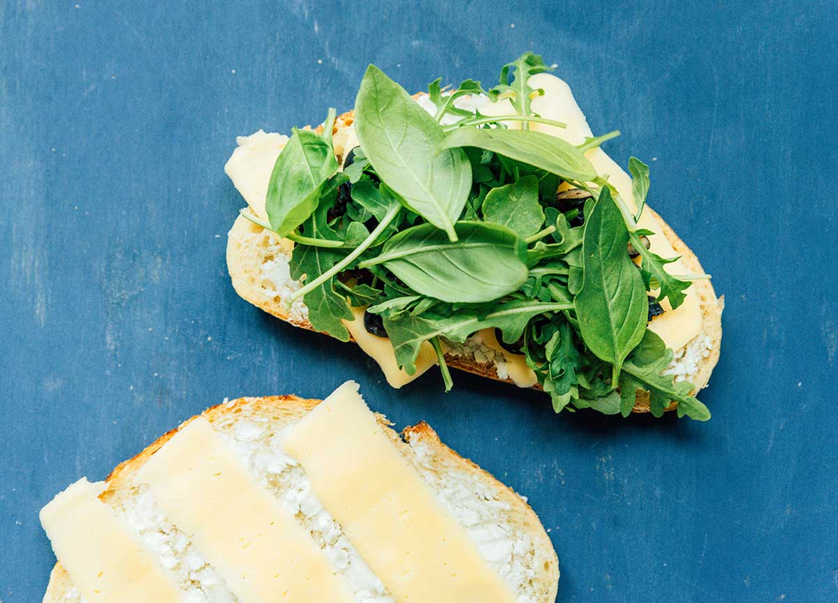 One piece of sourdough bread topped with goat cheese, a slice of mozzarella, halved blueberries, and arugula beside a second piece of bread topped with goat cheese and mozzarella