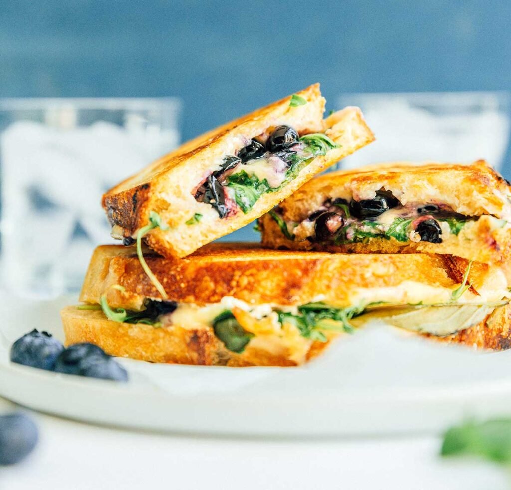 Two blueberry grilled cheese sandwiches on a plate, with one sandwich cut in half and stacked on top of the other