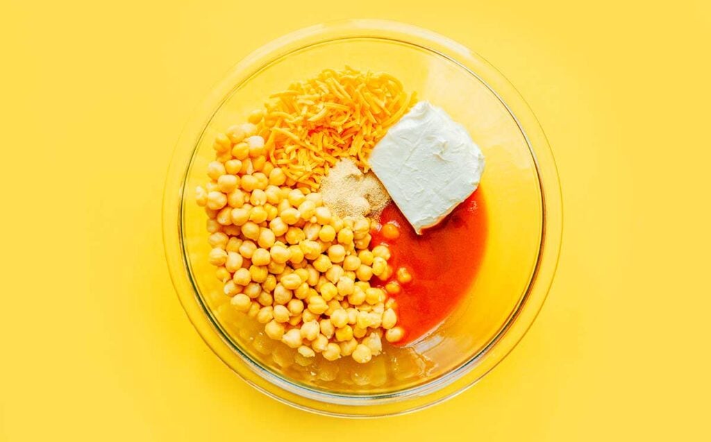 A glass bowl filled with chickpeas, garlic powder, cheddar cheese, cream cheese, and hot sauce