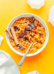 A bowl of rich pumpkin oatmeal, topped with crunchy pecans and fragrant cinnamon sticks, rests on a vibrant yellow background, flanked by two refreshing glasses of milk.