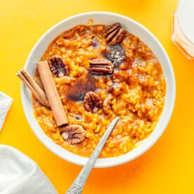 A bowl of rich pumpkin oatmeal, topped with crunchy pecans and fragrant cinnamon sticks, rests on a vibrant yellow background, flanked by two refreshing glasses of milk.