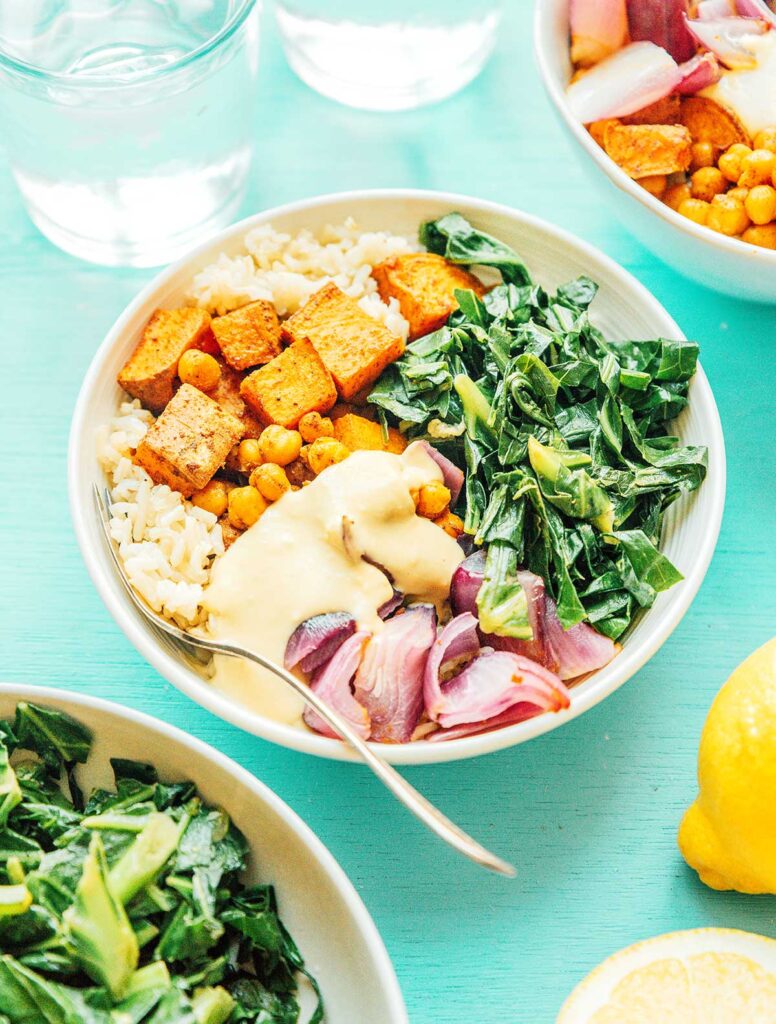 A southern style grain bowl filled with sweet potatoes, veggies, collard greens, red onion, and rice, and topped with a hummus and lemon juice sauce
