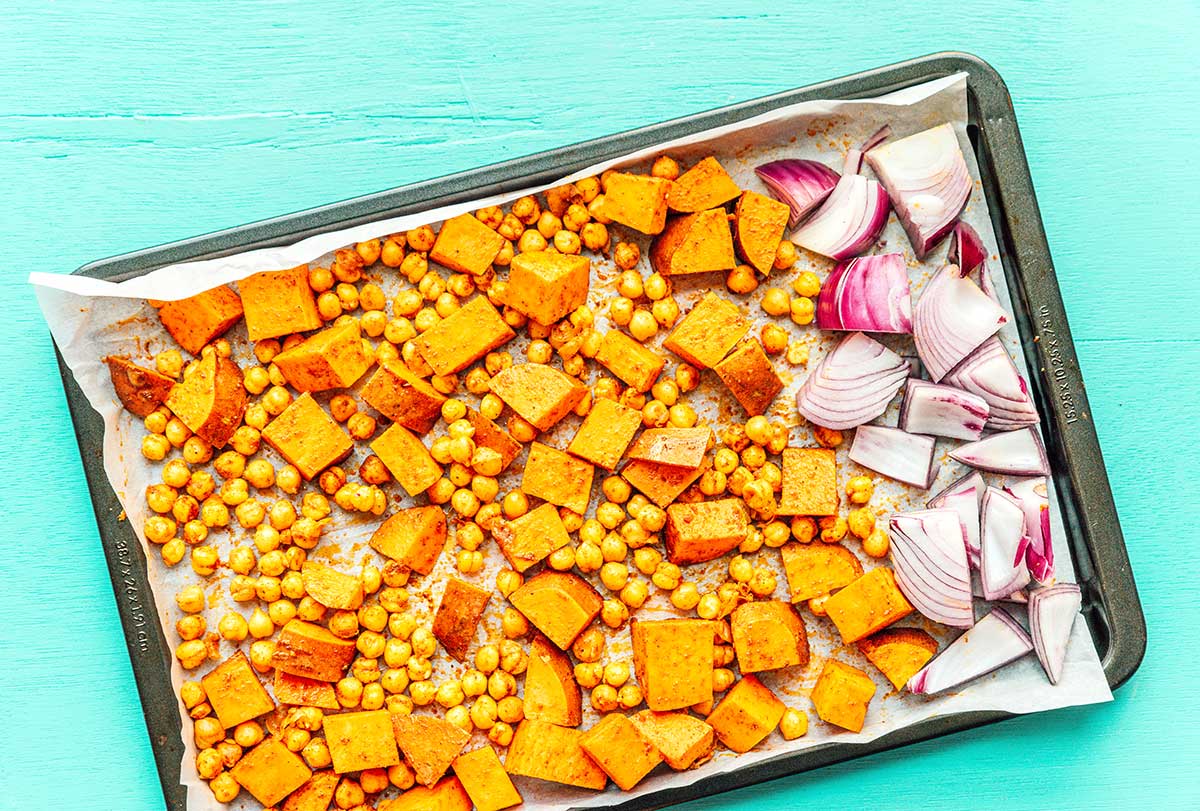 A baking tray filled with seasoned and chopped sweet potato, red onion, and a can of chickpeas