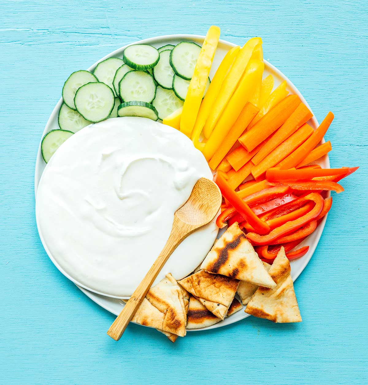 A bowl of whipped feta spread on a white plate surrounded by sliced cucumbers, sliced bell peppers, and pita bread