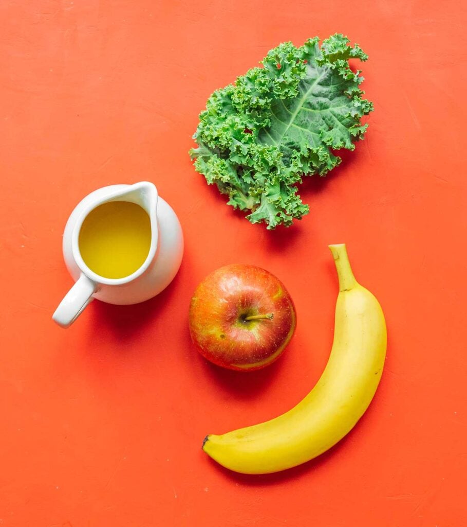 A piece of kale, a red apple, a banana, and a small pitcher with orange juice on a red orange background