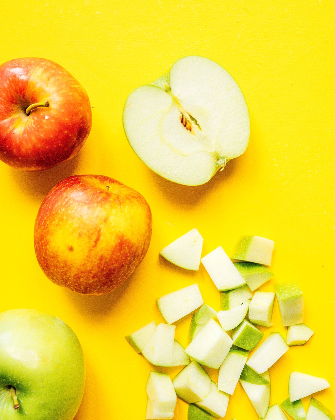 Three whole apples, an apple half, and diced apple pieces arranged on a yellow background