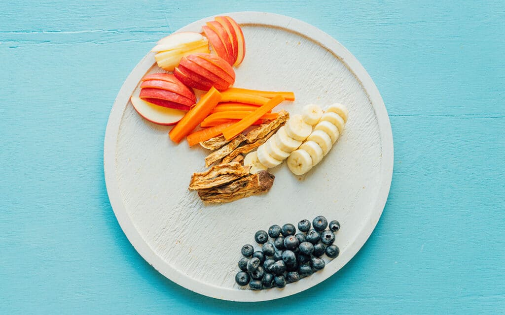 A barkuterie board with apples, carrots, dried sweet potatoes, bananas, and blueberries
