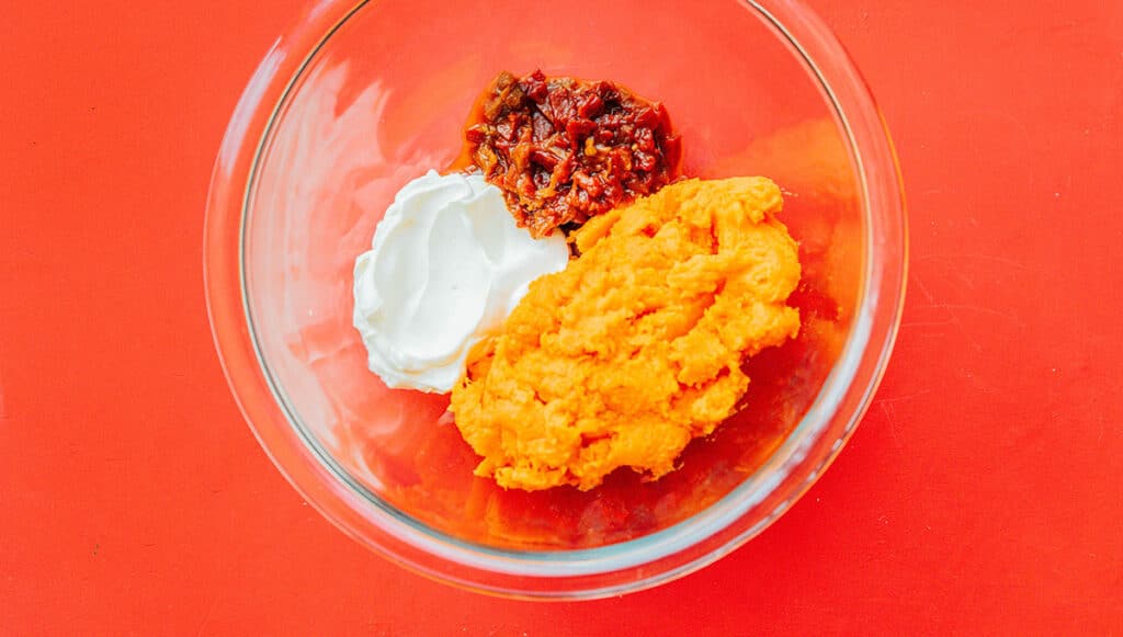 A clear glass bowl filled with sweet potato flesh, adobo peppers, and sour cream