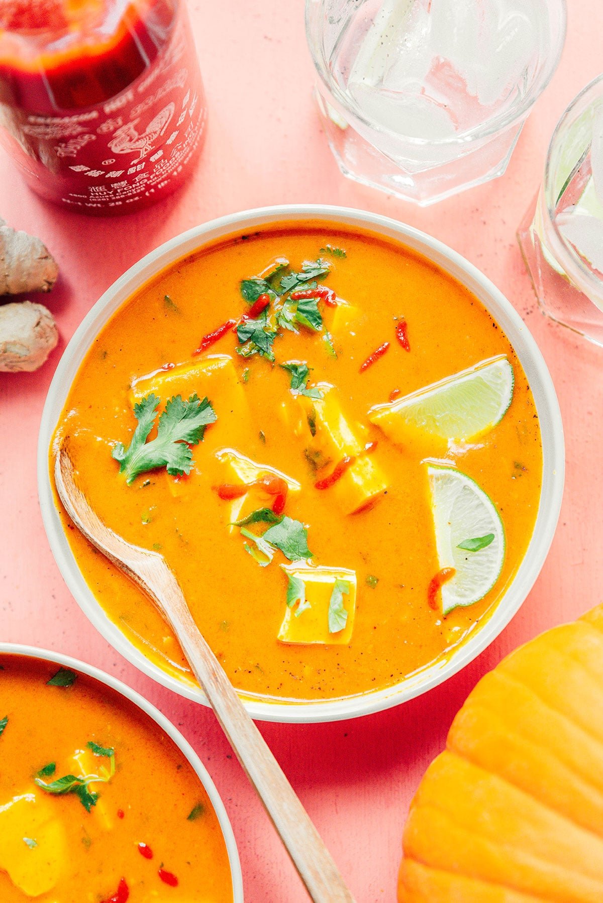 A white bowl filled with pumpkin curry garnished with cilantro, sriracha, and lime slices