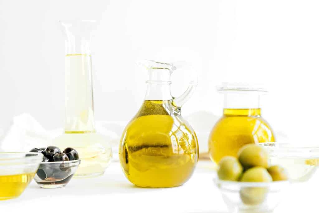 A glass handled jar containing olive oil surrounded by other olive oil containers and bowls of olives