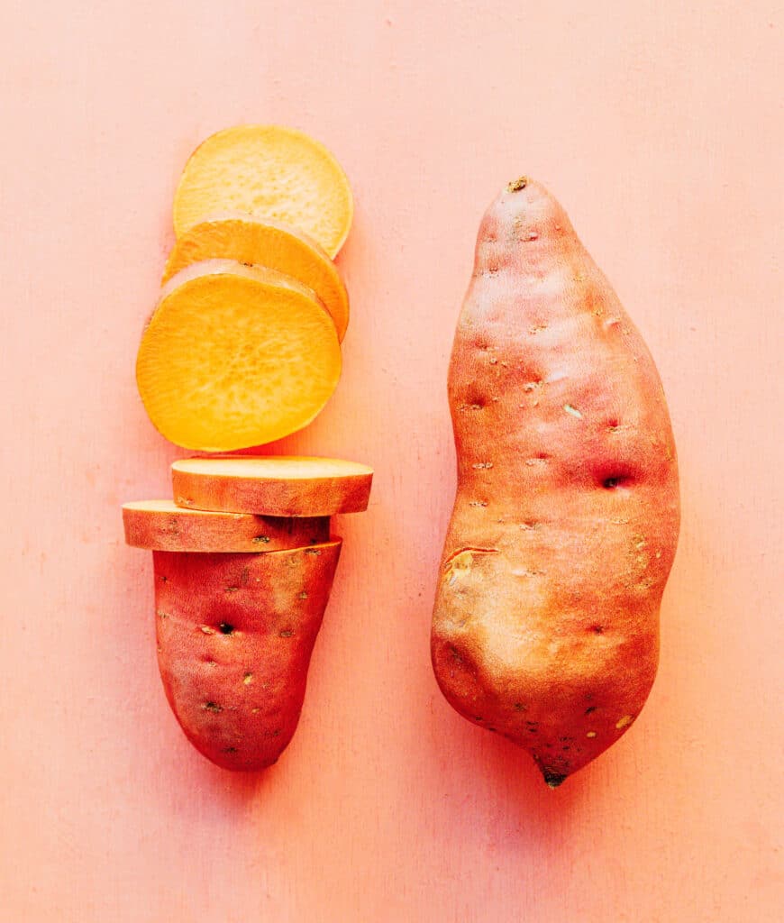 Two sweet potatoes sitting side by side, one of which is partially sliced to demonstrate the appropriate thickness of the rounds