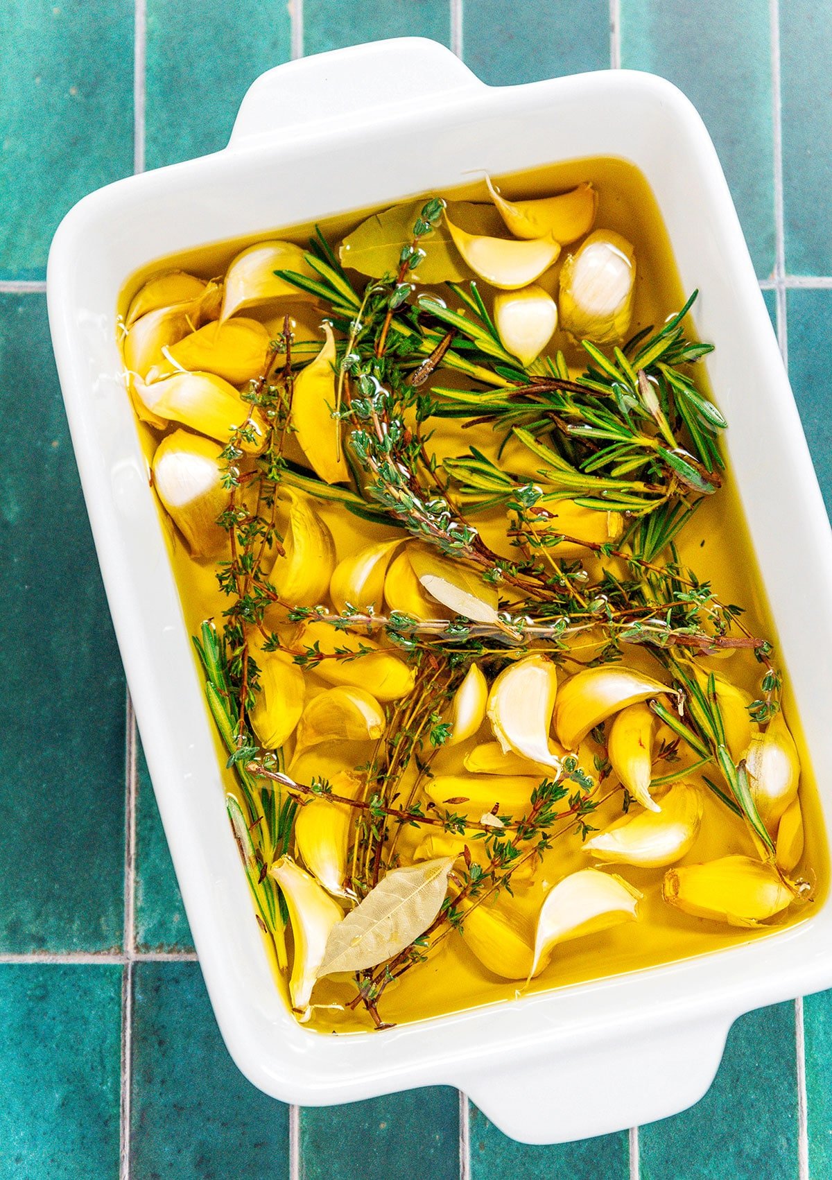 A casserole dish filled with garlic cloves soaking in olive oil and topped with fresh thyme, rosemary, and bay leaves