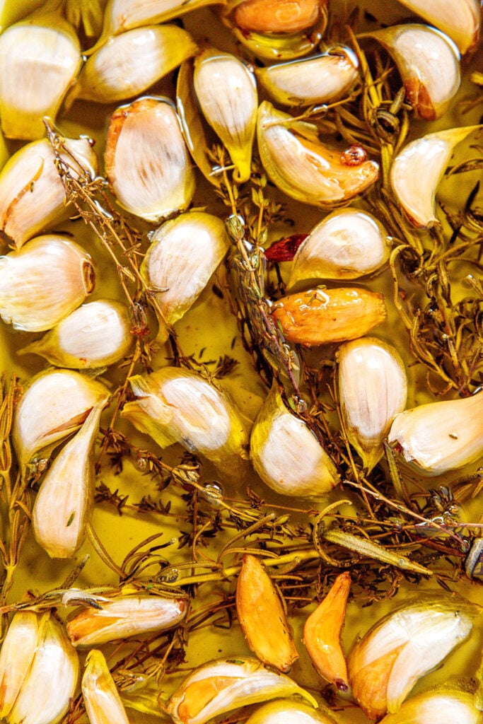 A close up view detailing the texture of cooked garlic cloves submerged in olive oil and topped with cooked herbs