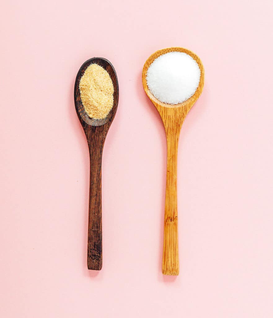 Two wooden spoons placed side by side on a pink background, the left holding garlic powder and the right holding salt