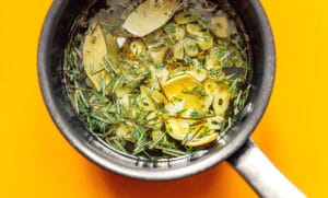 Saucepan with marinated olives, garlic, herbs, bay leaves, and oil on a bright orange surface.