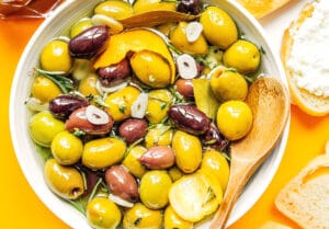 A bowl of marinated olives with herbs, garlic slices, and lemon zest, accompanied by a wooden spoon. Bread with cheese is visible at the edges.