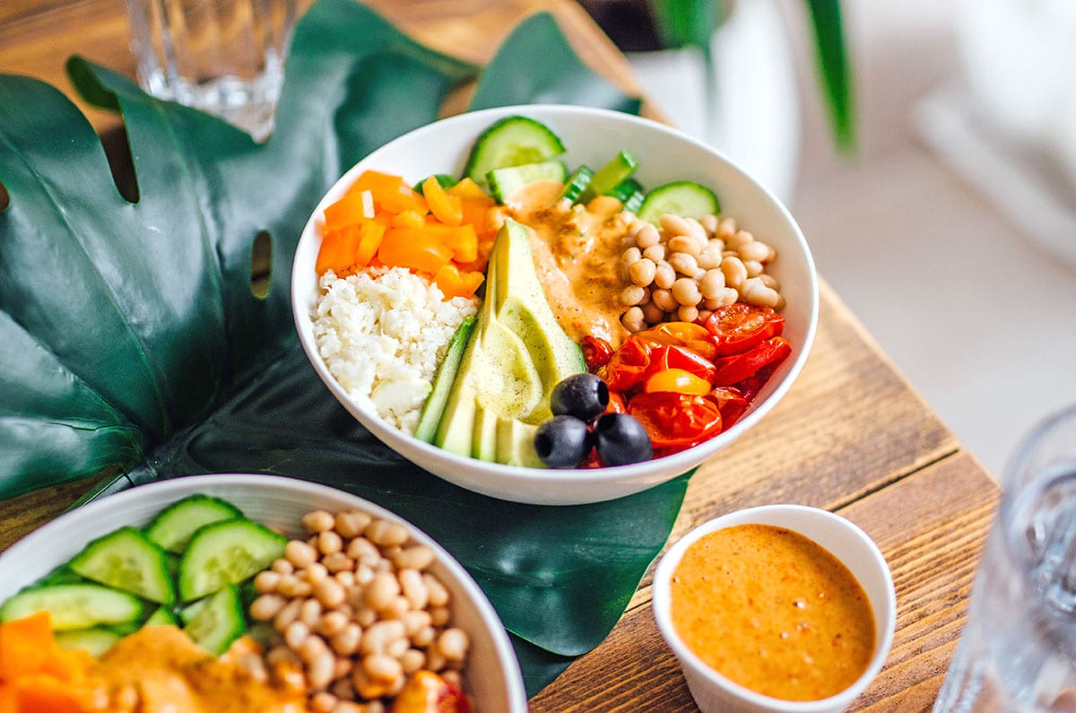 A Mediterranean romesco bowl made with grated cauliflower, diced bell pepper, roasted tomatoes, sliced avocado, sliced cucumber, white beans, and black olives and topped with a creamy romesco sauce