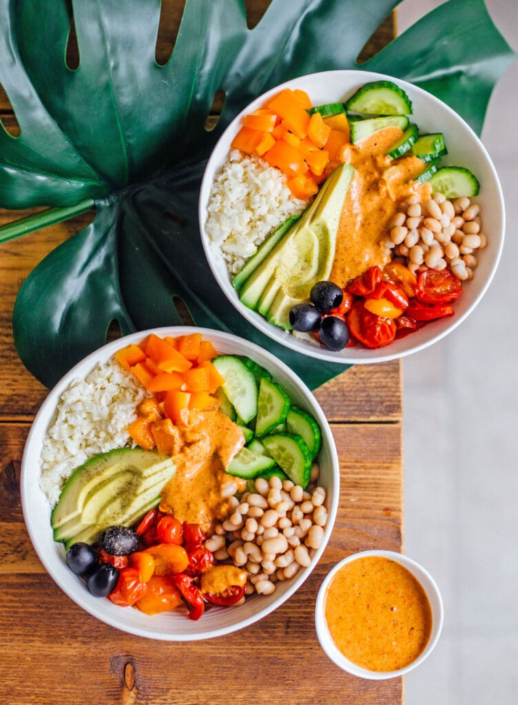 Two white bowls filled with a Mediterranean bowl recipe with ingredients like grated cauliflower, sliced avocado and cucumber, diced bell pepper, white beans, black olives, and roasted tomtaoes