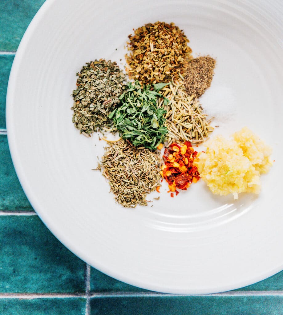 A white plate filled with spices including minced garlic, basil, thyme, oregano, rosemary, parsley, crushed red pepper flakes, salt, and pepper