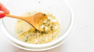 A wooden spoon mixing a creamy yellow sauce with herbs in a clear glass bowl, perfect for drizzling over vegan garlic bread.