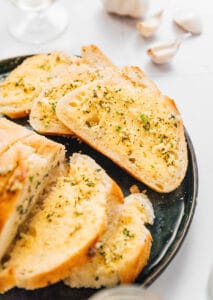 Slices of vegan garlic bread fragrant with herbs sit on a dark plate. In the background, a few garlic cloves are artfully displayed.