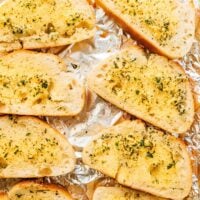 A tray of vegan garlic bread slices, topped generously with herbs, is artfully arranged on foil.