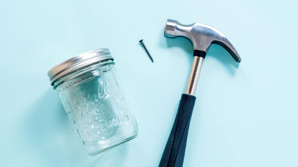 An empty mason jar, a nail, and a hammer placed on a blue background