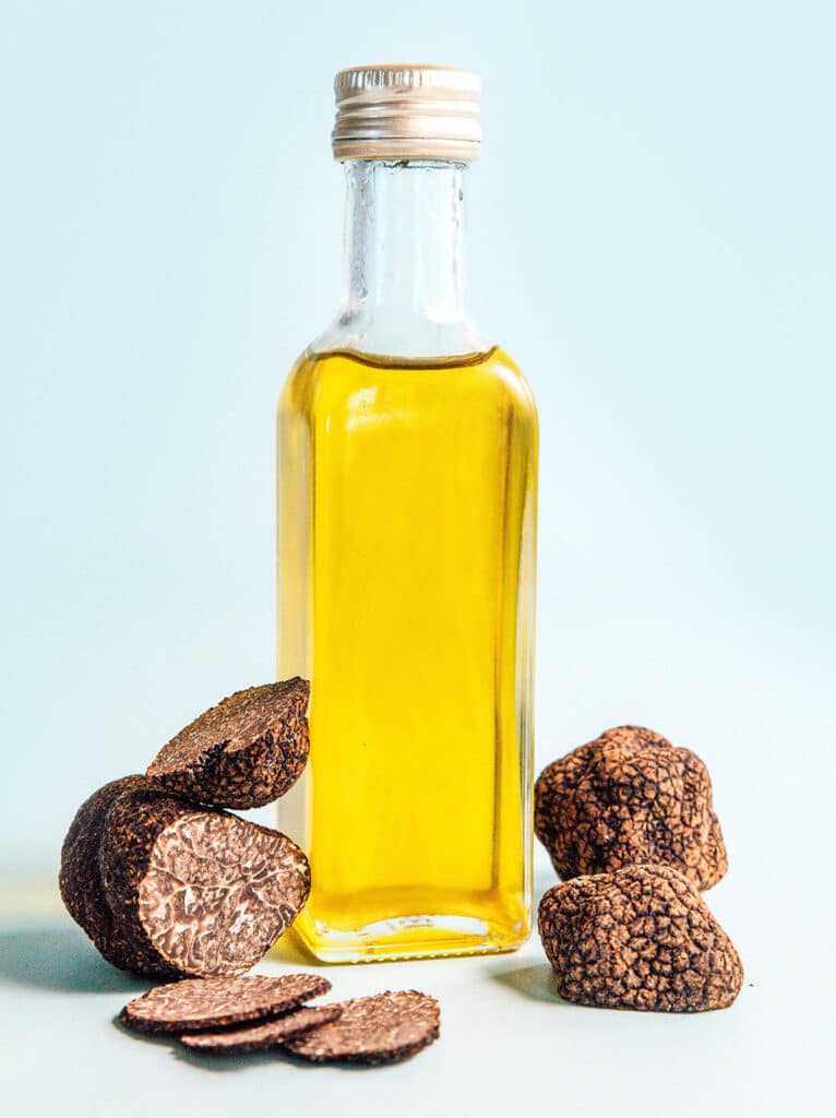 A glass jar filled with black truffle oil surrounded by whole and shaved black truffles
