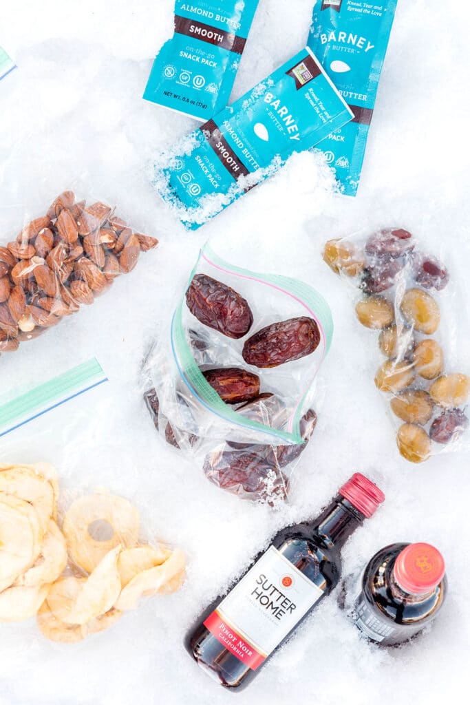 An arrangement of snacks lying on the snow including Barney Smooth Almond Butter, Sutter Home Pinot Noir, dates, olives, almonds, and dried fruit