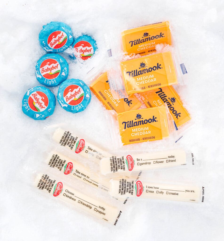 A selection of cheese laid out on the snow including 5 wheels of Light Babybel, 5 individually wrapped mini squares of Medium Cheddar Tillamook, and 5 mozzarella cheese sticks