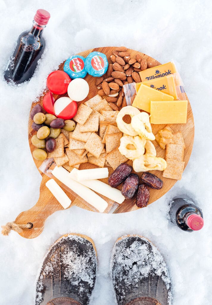 A wooden board on top of snow filled with an arrangement of cheese, crackers, dried fruit, and more, surrounded by a pair of snow covered boots and two mini bottles of wine