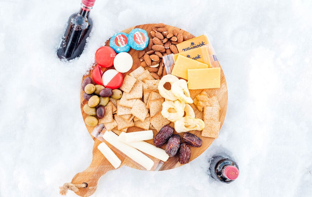 A wooden board on top of snow filled with an arrangement of cheese, crackers, dried fruit, and more, with a mini bottle of wine on either side