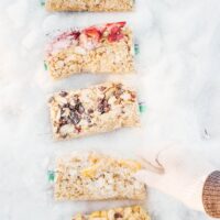 A gloved hand reaches towards five rectangular blocks of mixed grains and fruits, resembling oatmeal packs, arranged on the pristine snow.