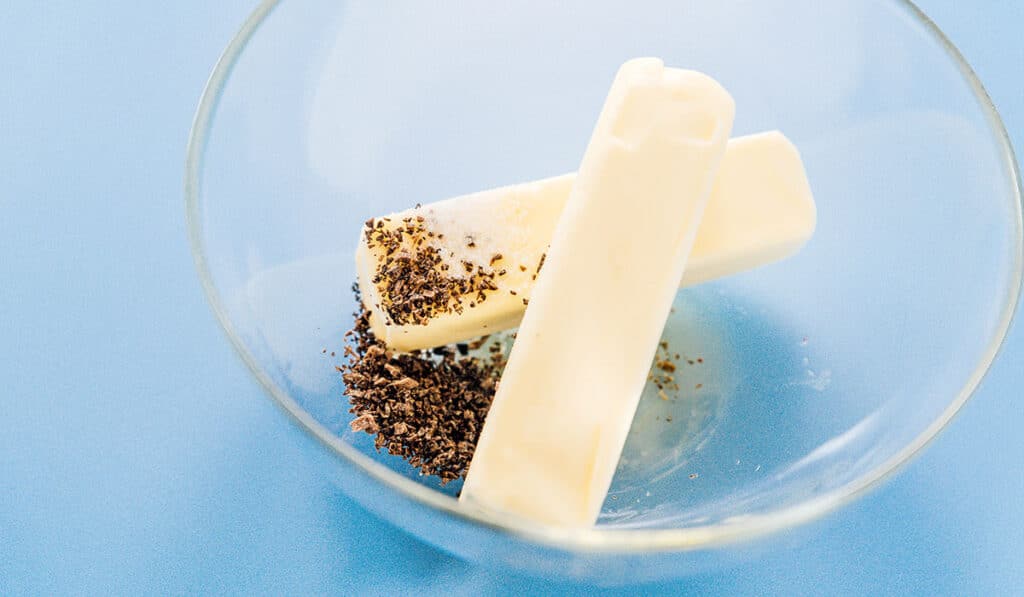 A clear glass bowl filled with two softened sticks of butter, truffle oil, finely chopped black truffle, and salt
