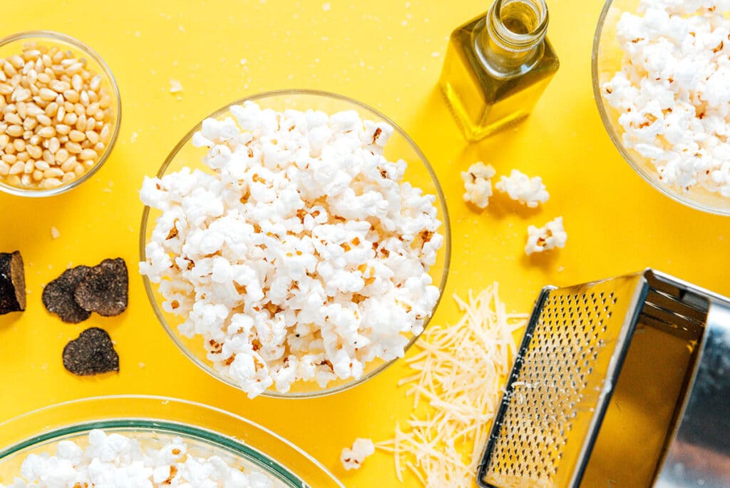 A bird's eye view of three glass bowls filled with homemade truffle popcorn, with various items scattered around: cheese grater, parmesan, kernels, truffles, and oil
