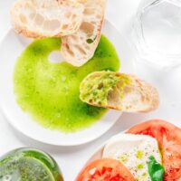 Slices of bread with green sauce, a hint of how to make basil oil, rest on a white plate. Nearby is a salad featuring tomatoes, mozzarella, and fresh basil. Accompanying the meal are glasses of water and a refreshing green drink.